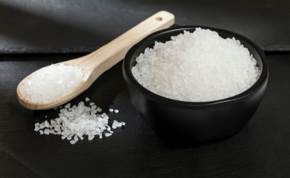 Eine Schwarze Schüssel und ein Holzlöffel gefüllt mit Salz liegen auf schwarzem Hintergrund